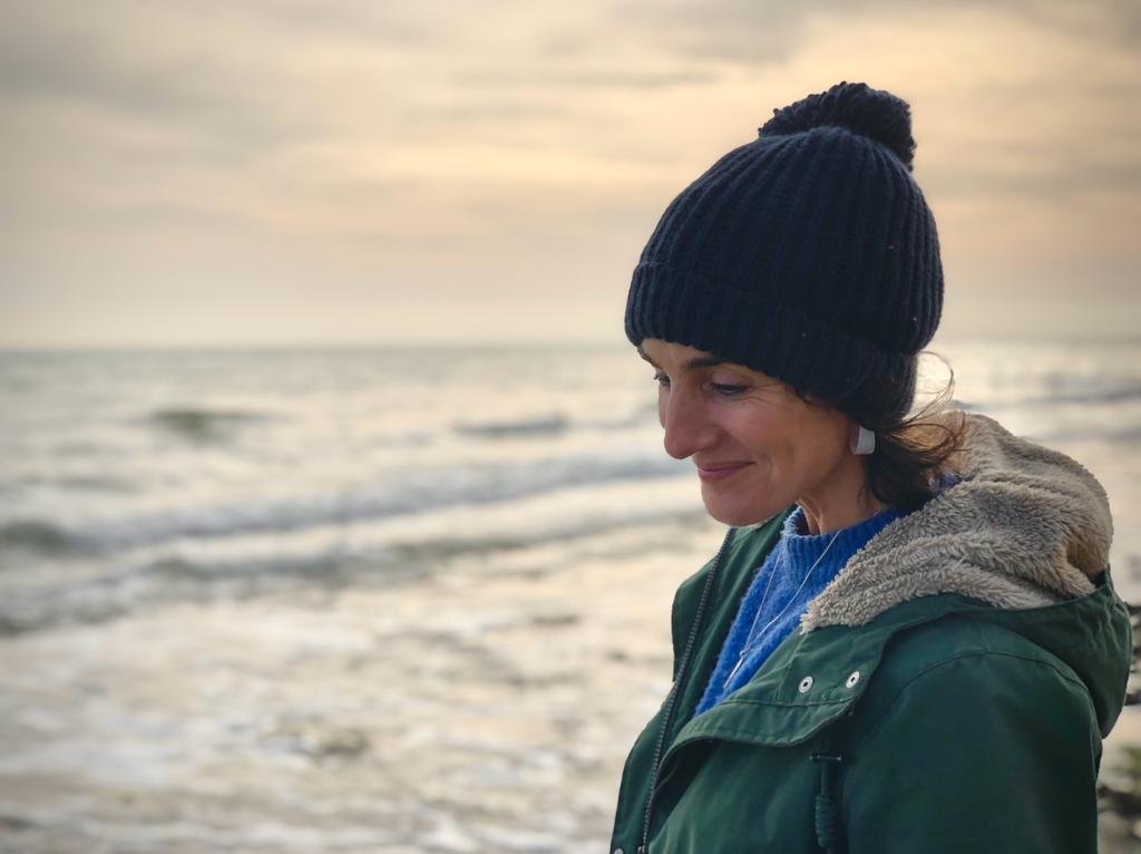 Xenia on a winter beach in a woollen hat and green coat, looking down and smiling