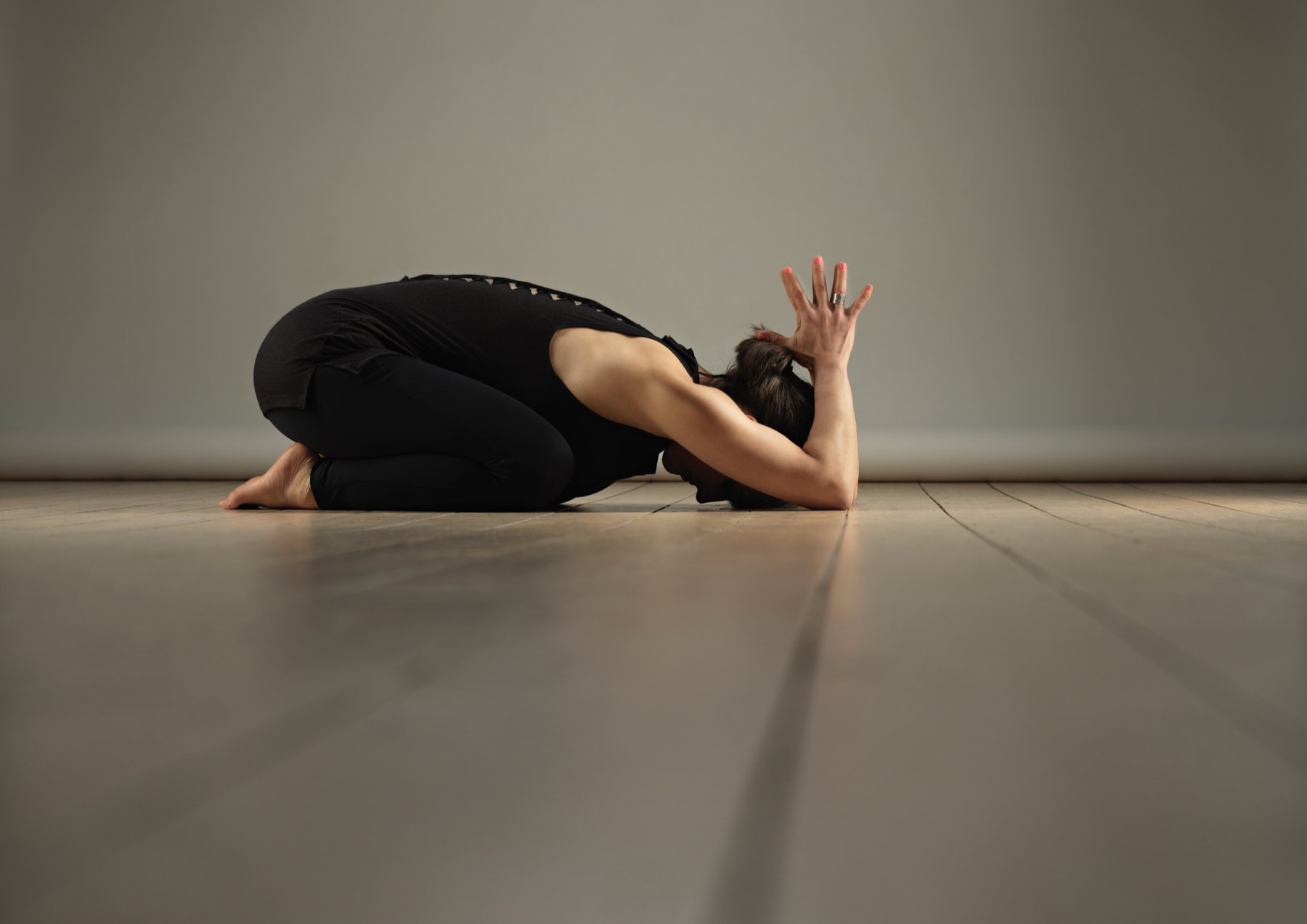Xenia resting in child's pose on a pale wooden floor, one hand raised behind the head